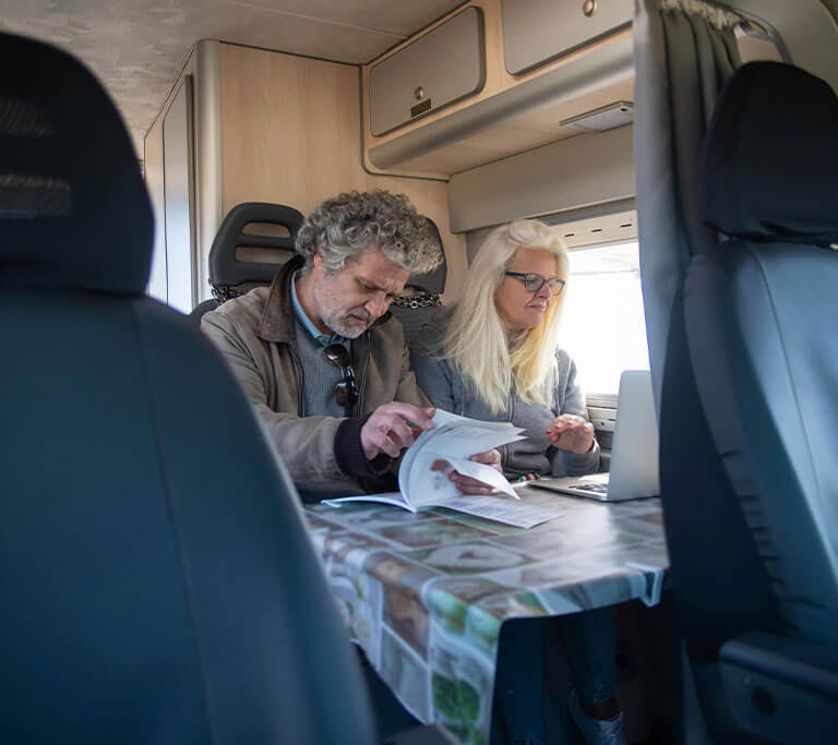 A couple sits at a table in an RV reading a book and using a laptop.