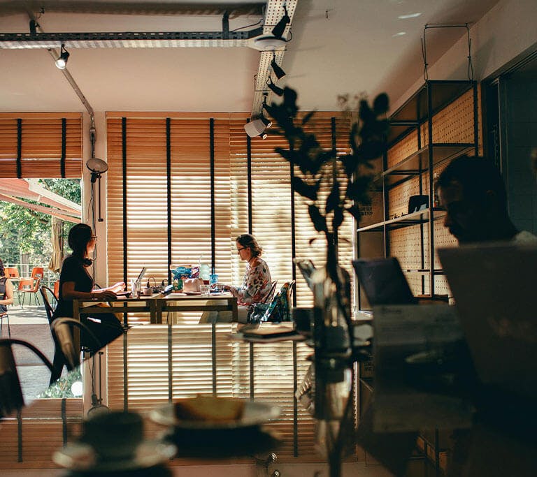 Several people work on laptops in a shared indoor space with large windows and natural light. Some are seated at communal tables, while others sit near the open entrance, suggesting a relaxed coworking or café environment.