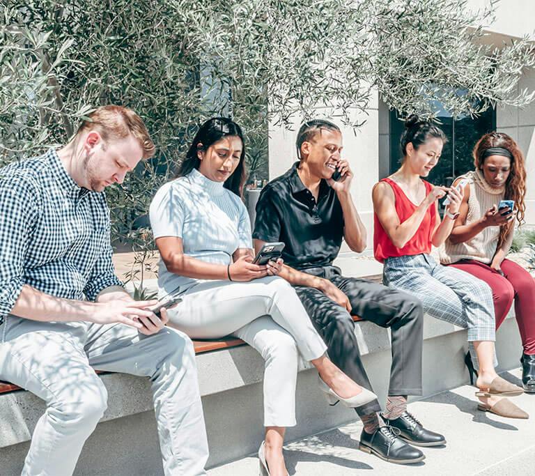 Group of people sitting on a bench outdoors, each using a smartphone, with trees and a building in the background.