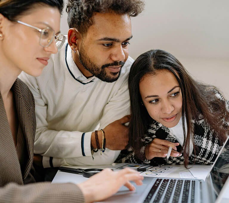Three people gathered around a laptop, looking intently at the screen while discussing a project. Papers with charts and designs are spread out on the table in front of them.