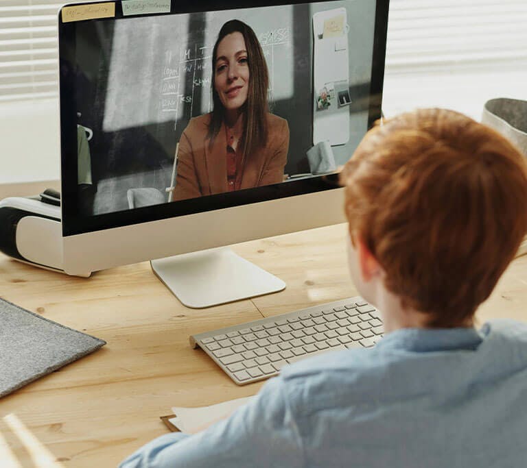 A child wearing a long-sleeve denim shirt sits at a Mac computer at a wooden desk while looking at a screen where their teacher, a person wearing a brown blazer, can be seen sitting in front of a chalkboard.