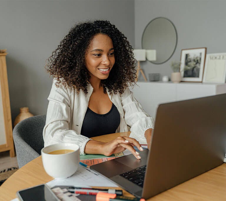 A person smiles while working on a laptop at a tidy desk with a coffee mug, highlighters and paper. The setting appears to be a cozy home office with framed art, plants and soft lighting in the background.