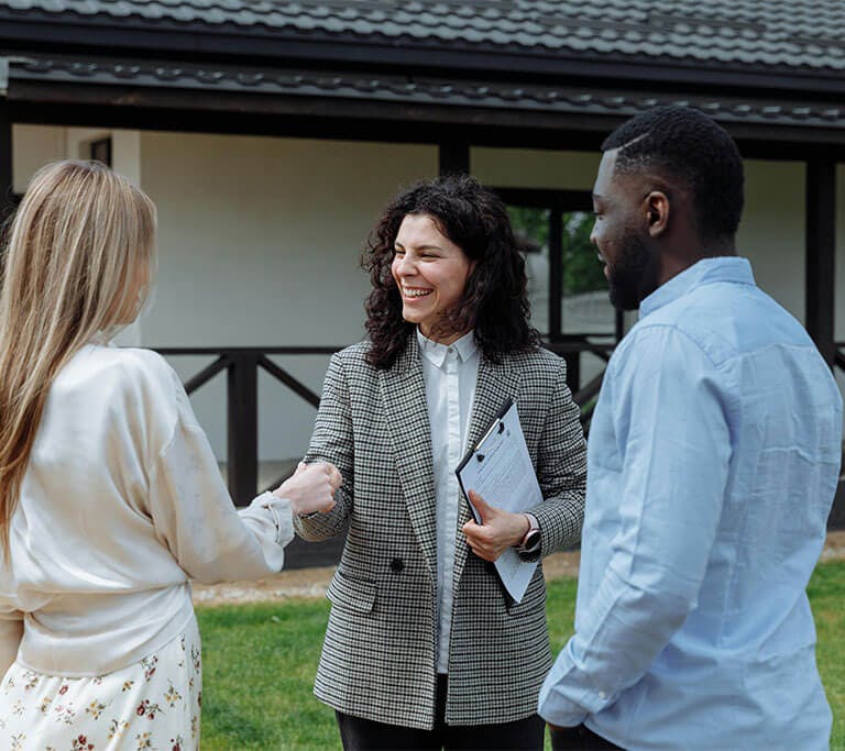 A person holding a clipboard and smiling while shaking hands with another person outdoors near a house. A third person stands beside them, suggesting a friendly meeting or discussion.