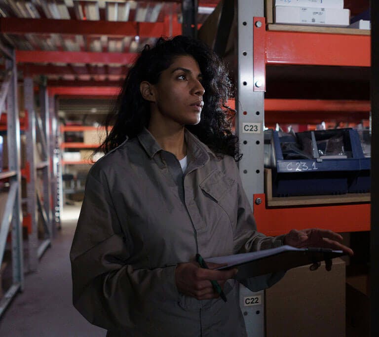 Person in work coveralls stands in a dim warehouse aisle holding a clipboard and pen, looking toward shelves filled with labeled boxes and bins.