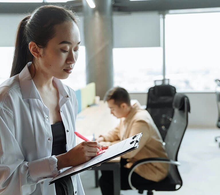 A person standing and writing on a clipboard with a pen in a modern office setting, while another person works at a desk in the background.