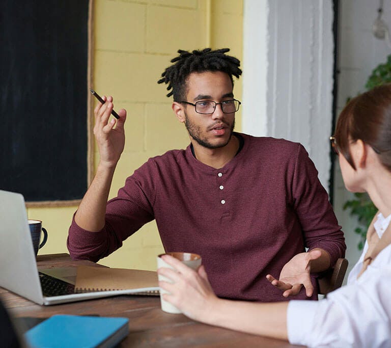 Two people sit at a table discussing something while looking at a laptop, with one person gesturing with a pen and one holding a coffee mug.