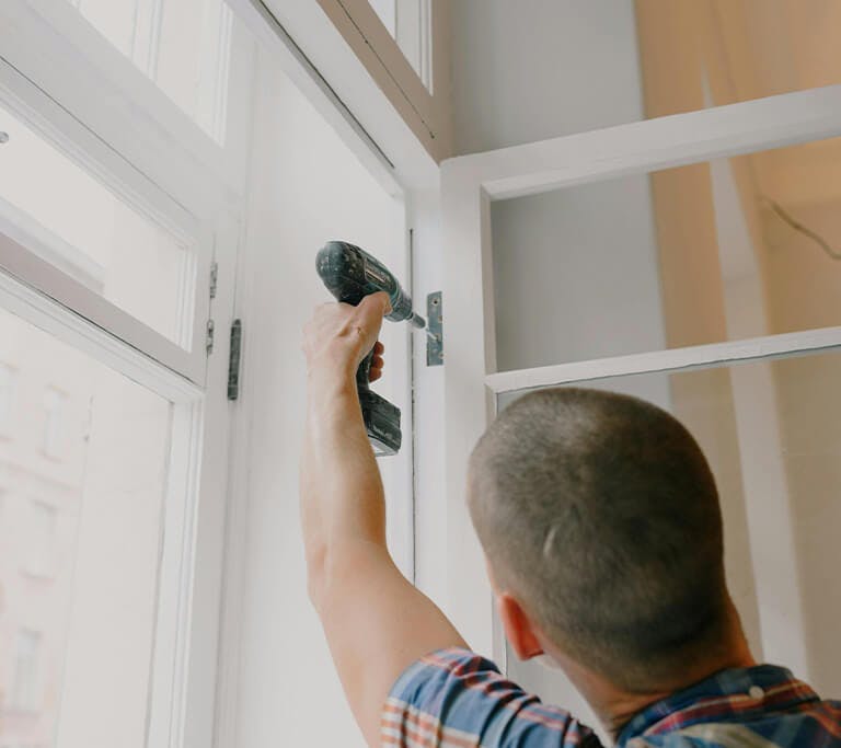 A person using a power drill to install or repair a window frame inside a bright room, with natural light coming through large windows.