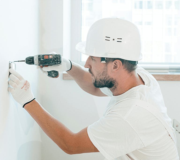 A person wearing a white hard hat, gloves, and a white shirt uses a power drill to install or secure a fixture to a white wall. A window with natural light and tools on the windowsill are visible in the background.