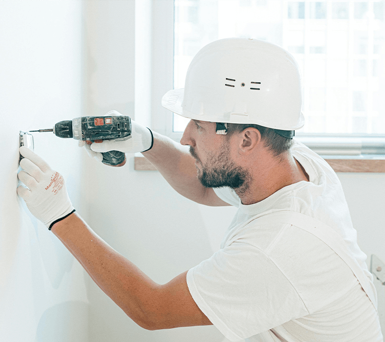 A person wearing a white hard hat, gloves, and a white shirt uses a power drill to install or secure a fixture to a white wall. A window with natural light and tools on the windowsill are visible in the background.