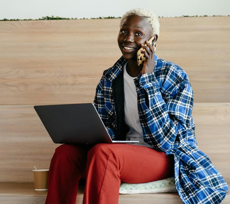 A person wearing a checkered blue button-up shirt and red pants smiles while talking on the phone and holding a laptop.