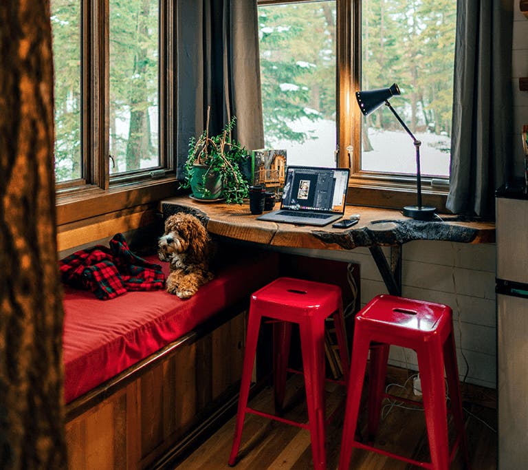 A small brown dog sits on a red-cushioned bench, part-way under a wooden table that has an open laptop computer sitting on it, beneath a flatscreen streaming TV that’s mounted to the wall above inside of a contemporary tiny home in a snowy, rural area.