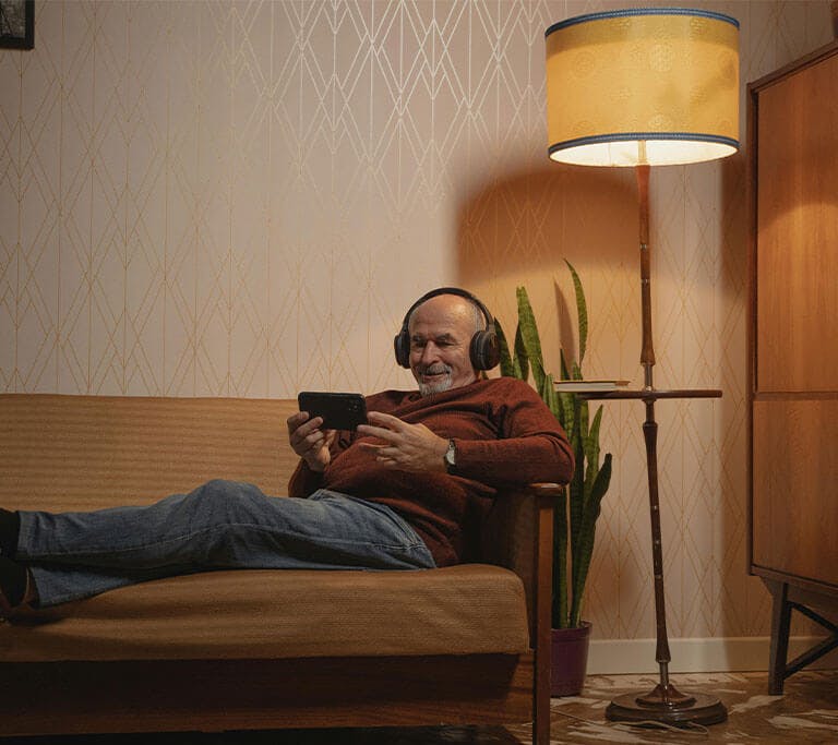 A person relaxes on a couch wearing headphones and streaming a video on a smartphone in a softly lit living room.
