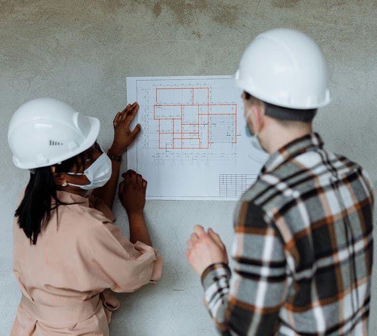 Two people wearing safety helmets and face masks reviewing a set of building plans pinned to a wall. One person points to the blueprint while the other observes, suggesting collaboration on a construction or architectural project.