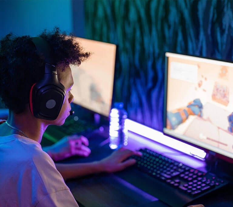 Two gamer friends sit side-by-side in a room with purple lights and play video games on desktop computers.
