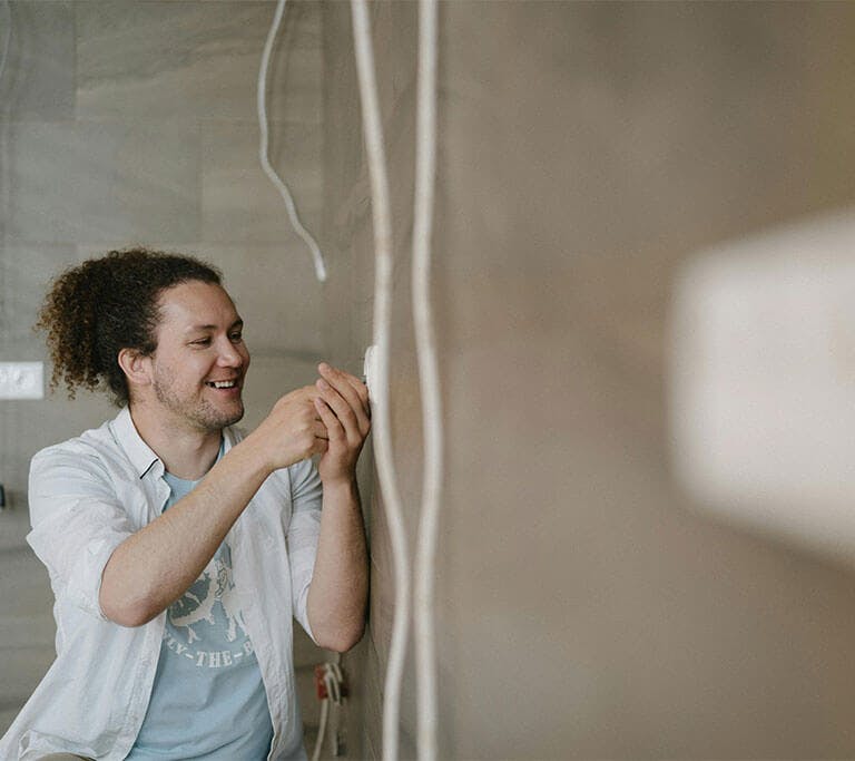 A person with curly hair wearing a white button-up shirt and a blue T-shirt underneath smiles as they install a smart thermostat into their wall.
