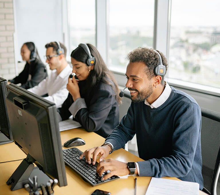 A group of customer service representatives wearing headsets work at desktop computers in a modern office with large windows. One person in the foreground is smiling while typing, suggesting a positive interaction.