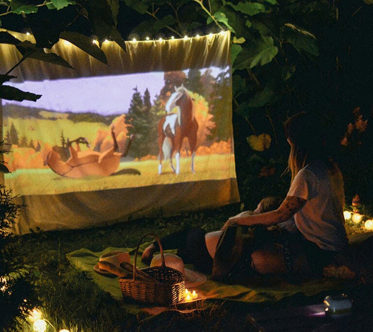 Two people sit on a blanket outside at night in a space dimly lit by string lights as they watch an animated film featuring two horses on a projector screen.