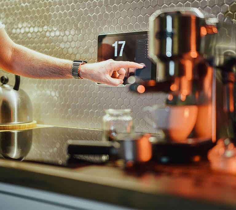 A person stands in a modern kitchen next to a smart coffee machine and uses their finger to navigate the device’s smart display.