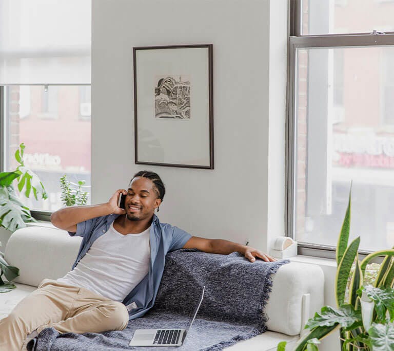 A person in a modern room with white walls and houseplant plants smiles as they talk on the phone while sitting on a white couch next to an open laptop.