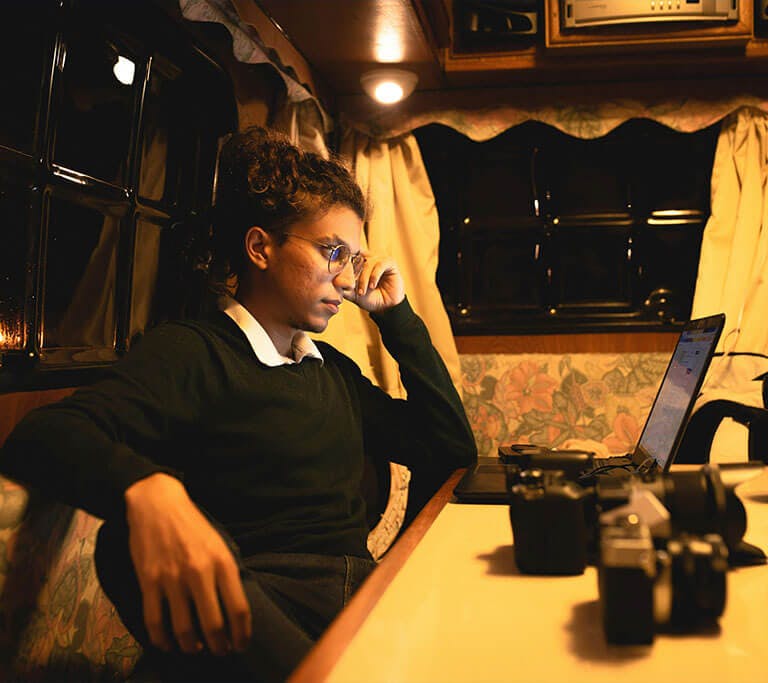 A person sitting inside a cozy camper van, working on a laptop at a table with several camera lenses and equipment, illuminated by warm indoor lighting