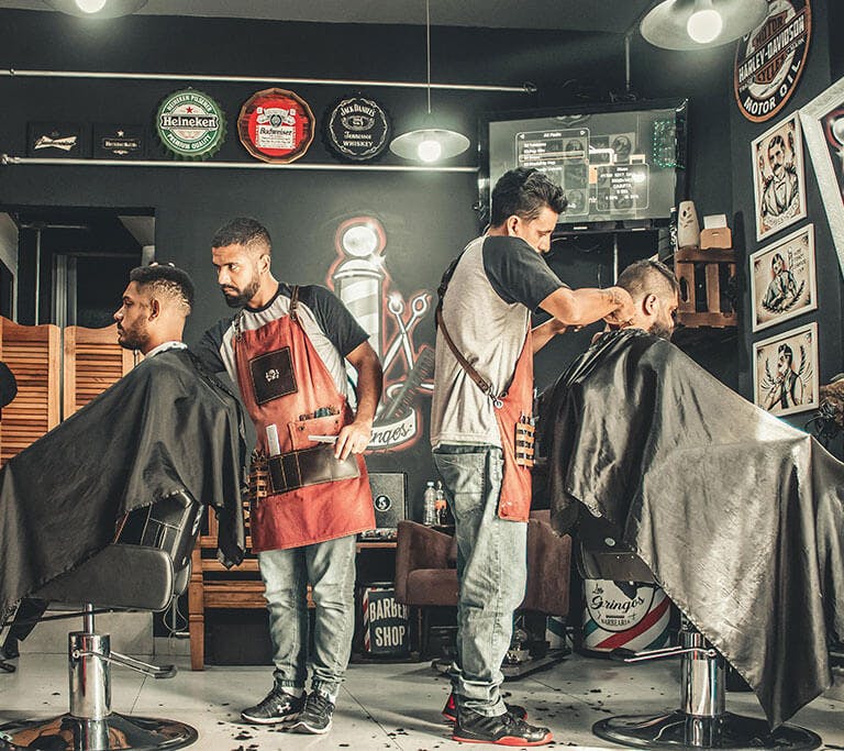 Two barbers cutting clients’ hair in a stylish barbershop decorated with vintage signs, framed artwork, and grooming tools. The atmosphere appears relaxed and professional, with each customer seated in a barber's chair under bright lighting.