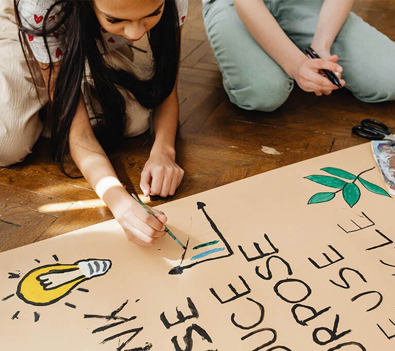 Two people sit on a wooden floor painting a large poster that shows a lightbulb, a plant and the words “REDUCE,” “REUSE,” and “RECYCLE.”