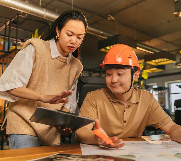 Two people reviewing documents together at a desk in a modern office, with one holding a tablet and the other wearing an orange safety helmet and gesturing toward a paper with a highlighter.