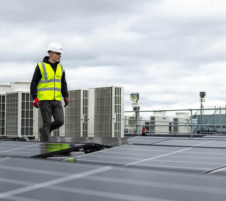A worker wearing a safety vest and hard hat walks across a rooftop solar panel installation, with HVAC equipment visible in the background.