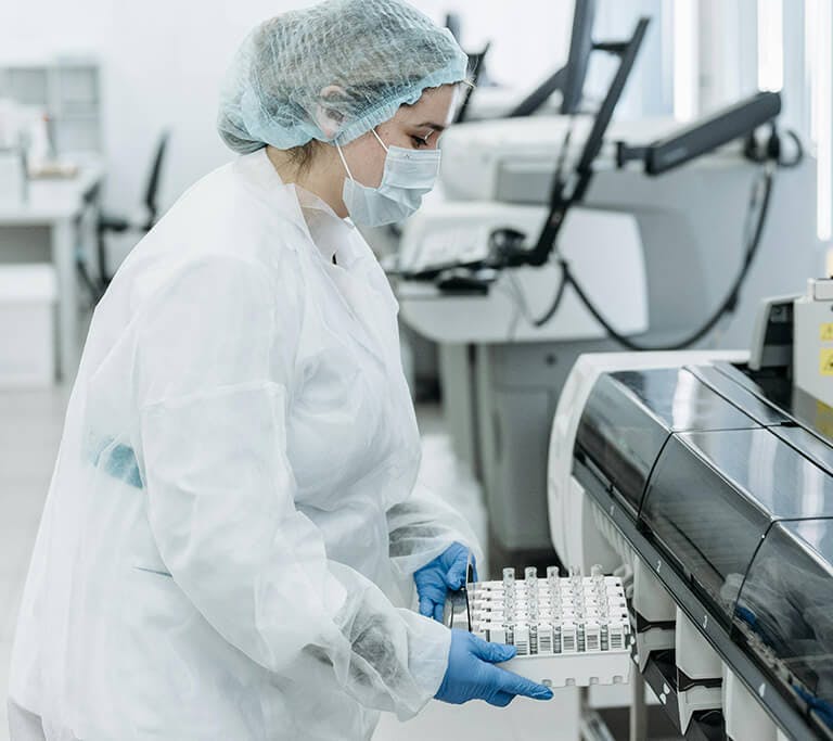 A laboratory technician wearing protective gear, including a mask, gloves, and hairnet, operates a machine while loading test tubes or samples inside a modern lab.