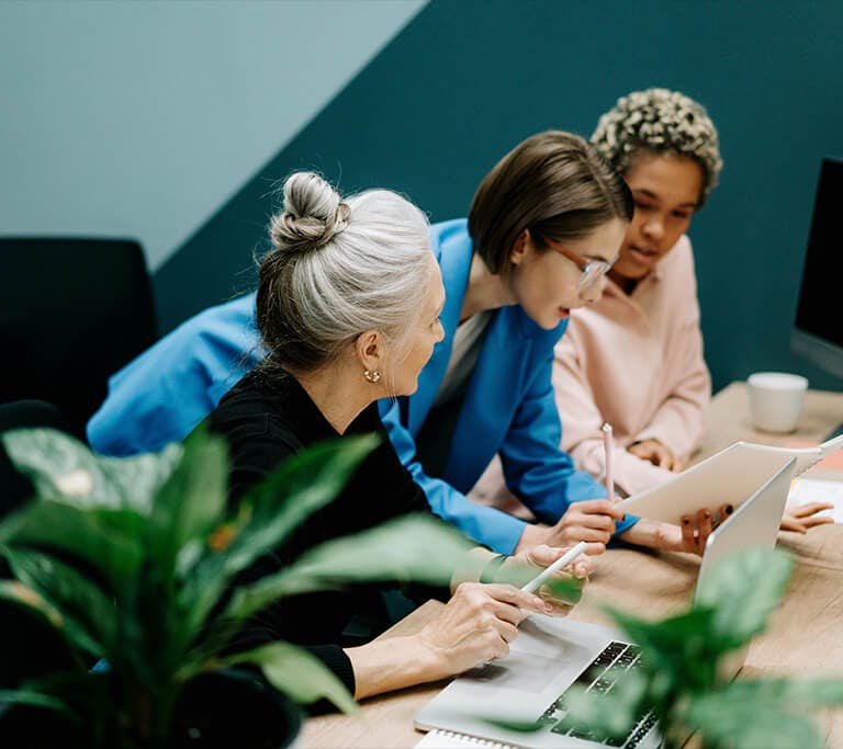 Three people collaborate at a desk, looking at a tablet and taking notes while working on laptops in a modern office setting with plants in the foreground.