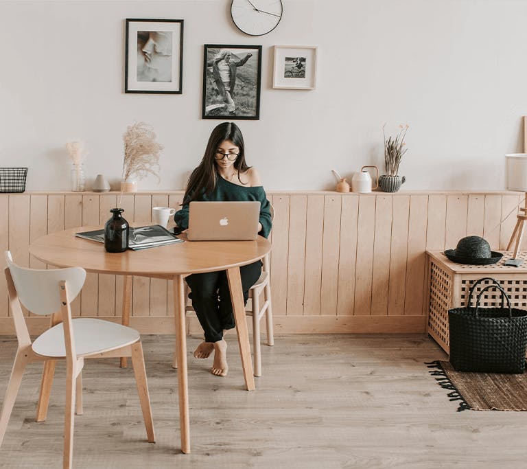 A person sits barefoot at a round wooden table working on a laptop in a cozy, well-lit room with minimalist decor. The space features light wood paneling, modern artwork on the wall, a rug, and various decorative items like vases, a hat, and a basket.