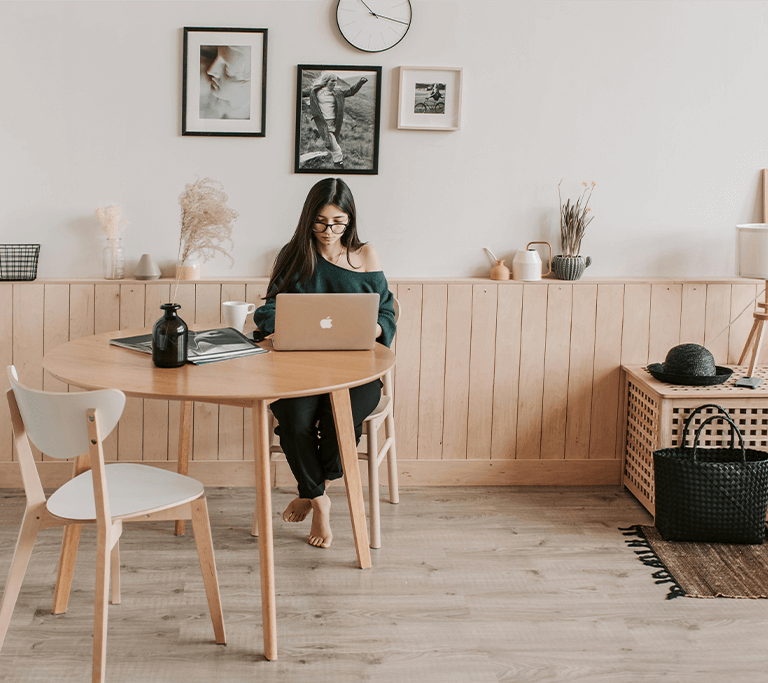 A person sits barefoot at a round wooden table working on a laptop in a cozy, well-lit room with minimalist decor. The space features light wood paneling, modern artwork on the wall, a rug, and various decorative items like vases, a hat, and a basket.
