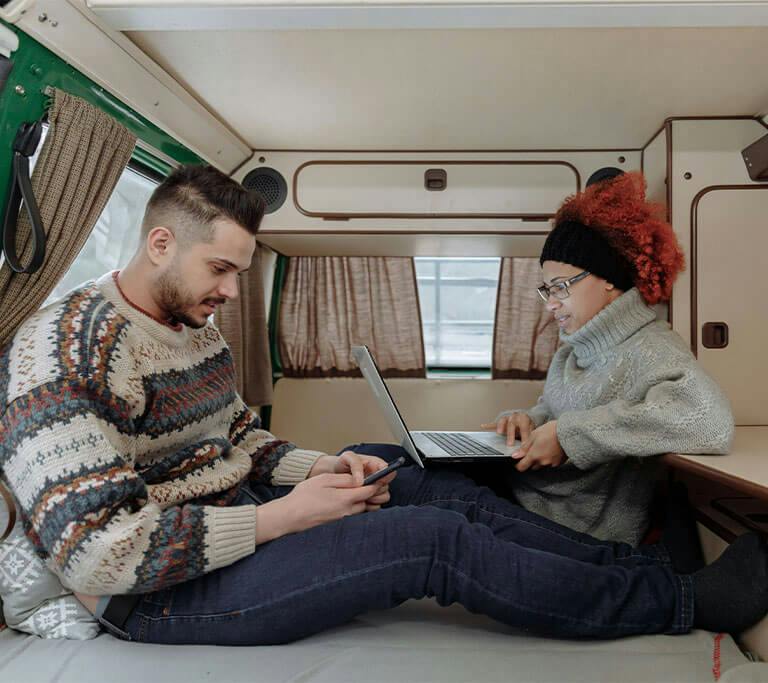 Two people sit inside a camper van. One is using a smartphone and the other is working on a laptop.