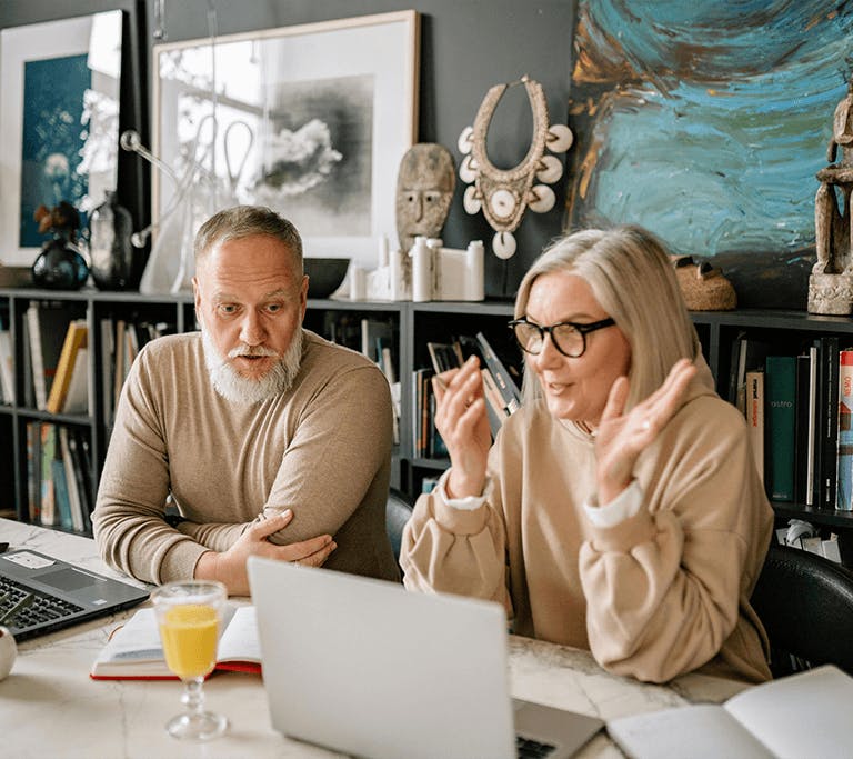 A silver-haired couple sits at their open laptop computers while having a discussion, each with a book open near them on a marble table in front of a wide bookshelf with artwork and sculptures lining the top.