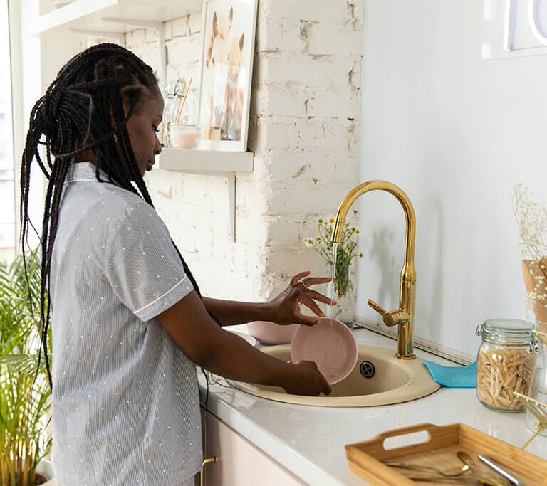 A person with long braided hair washes a pink bowl in a beige sink with a golden faucet on a sunny day.
