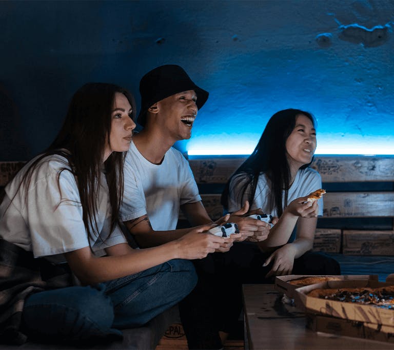 Three people sit on a couch in a dimly lit room with a blue LED light behind them, playing video games and sharing pizza. Two are holding game controllers and focused on the screen, while one is smiling and holding a slice of pizza. The atmosphere is casu