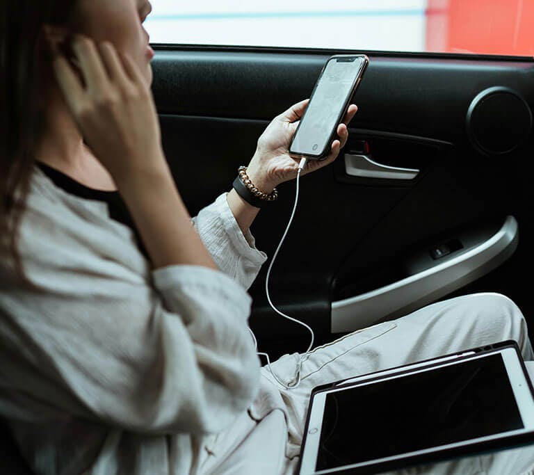 A person rides in the back seat of a car streaming music on a smartphone with wired earbuds, with a tablet resting on their lap.
