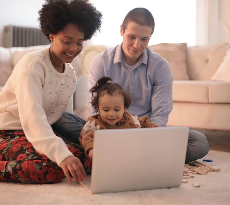 Two parents and a child sit on a white, soft rug on the floor while using a laptop and smiling.