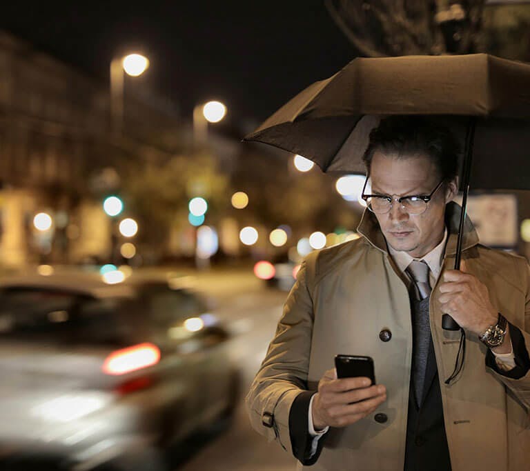 A person wearing glasses and a beige coat holds an umbrella while looking at his phone in a city at night.