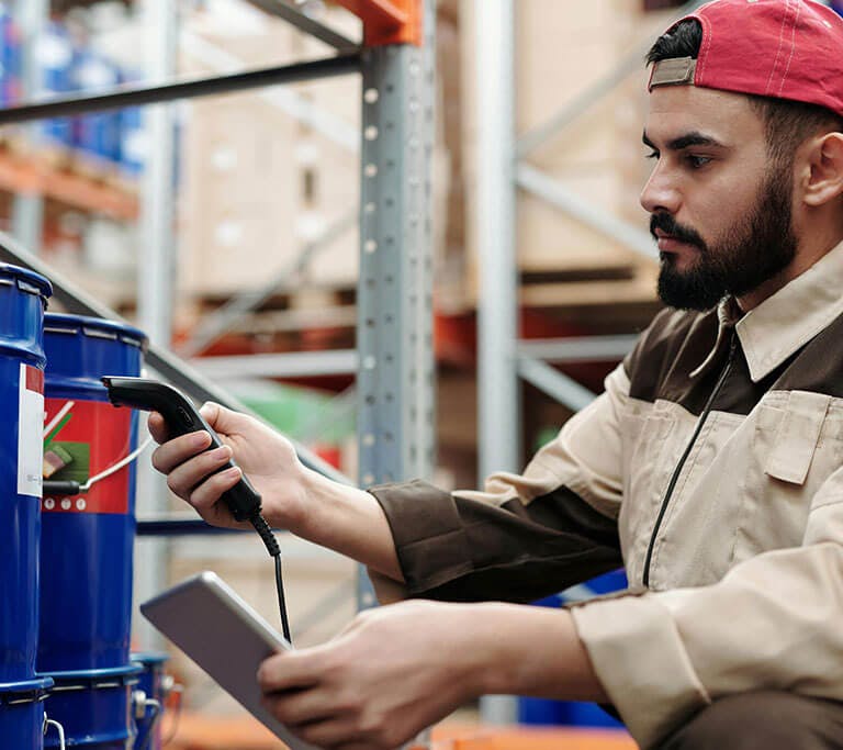 A person wearing a work uniform and backward cap scans a barcode on a blue container in a warehouse. They are holding a handheld scanner in one hand and a tablet in the other, suggesting they are managing inventory or performing a stock check.