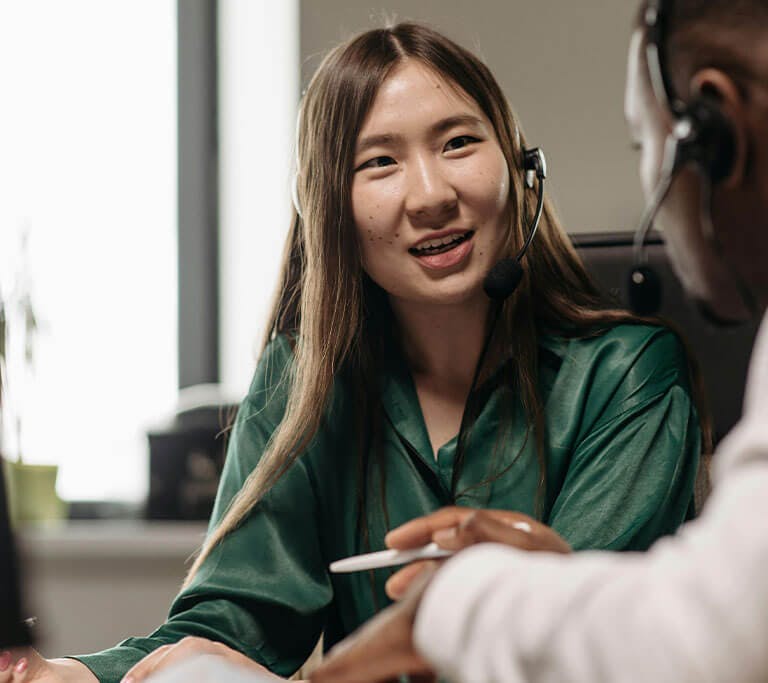 Two people wearing headsets are seated at a desk, engaged in a conversation. One person is smiling while speaking, and the other is listening attentively while holding a pen. A computer monitor is visible in the foreground.