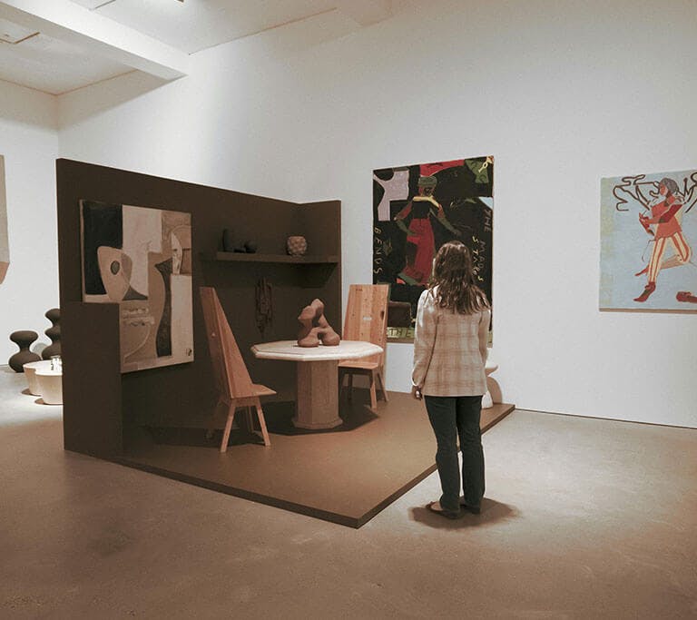 A person standing in an art gallery observing modern paintings and sculptures under motion-activated lights. The room features minimalist décor with abstract artworks on the walls and wooden furniture arranged as part of an exhibit.
