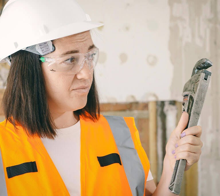 Person wearing an orange safety vest, safety glasses and a hard hat holding a plumbing tool in a white room.