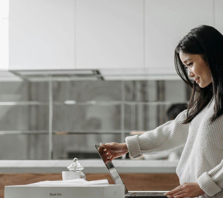 A person wearing a cream long-sleeve ribbed sweater smiles at a laptop screen in a minimalist kitchen with white, silver and light wood accents.