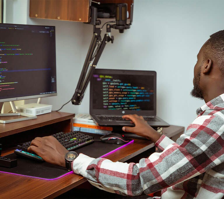 A developer wearing a checkered, long-sleeve shirt sits at a desk with a backlit keyboard and desktop as they review code on a laptop and display.