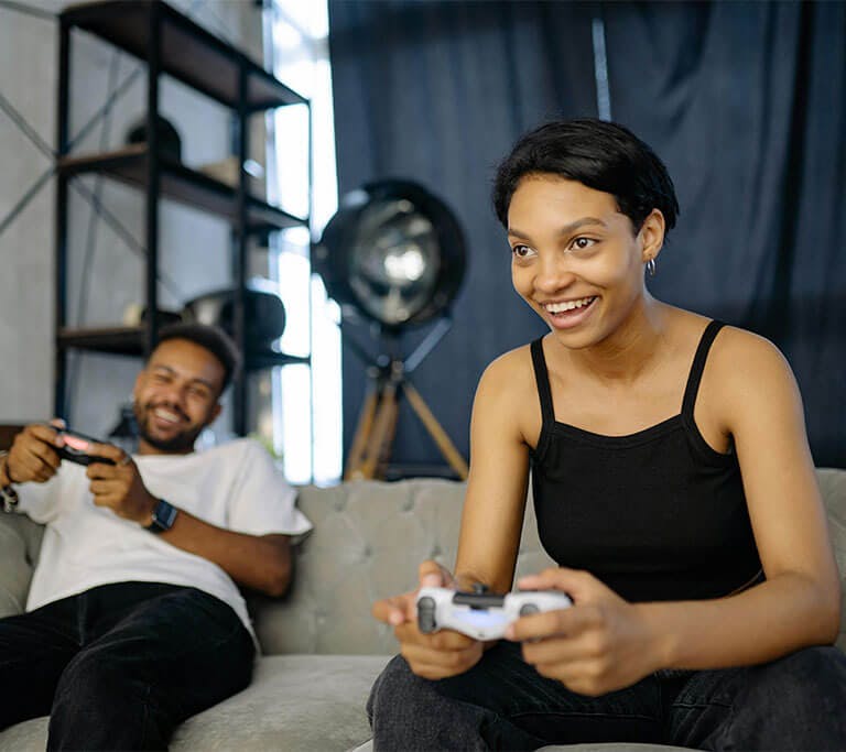 Two people sit on a couch playing video games together with wireless controllers. One person in the foreground smiles while focusing on the game, while the other lounges and laughs in the background. A large light and shelving unit are visible behind them
