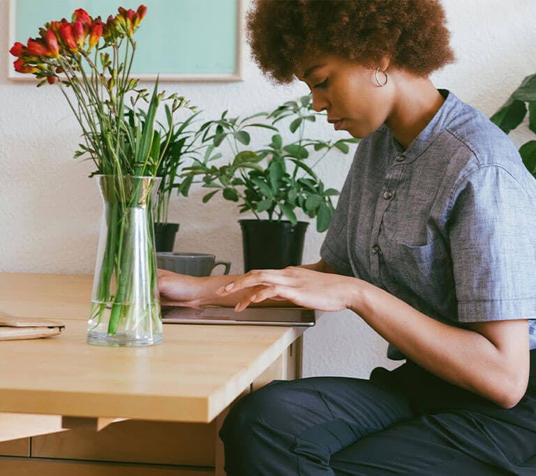 A person uses a tablet at a wooden desk in a room with houseplants and flowers in a vase.