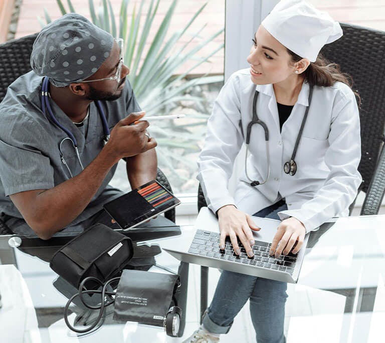 Two medical professionals sitting at a glass table, discussing something while one types on a laptop, and the other holds a tablet and pen, with a stethoscope and blood pressure monitor on the table.