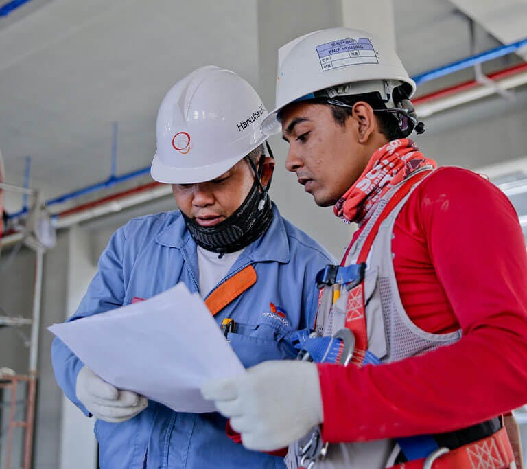 Two construction workers wearing safety helmets and protective gear reviewing documents at a job site. Scaffolding and building materials are visible in the background, indicating an active construction environment.