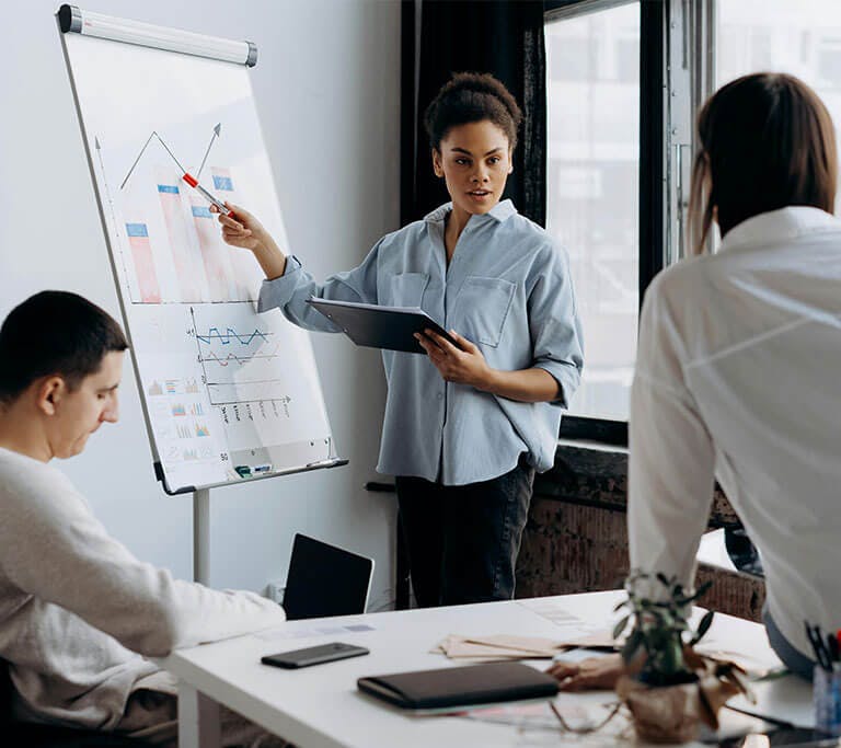 Person standing by a flip chart points to graphs during a presentation, while two others sit and lean at a desk listening in a bright office.